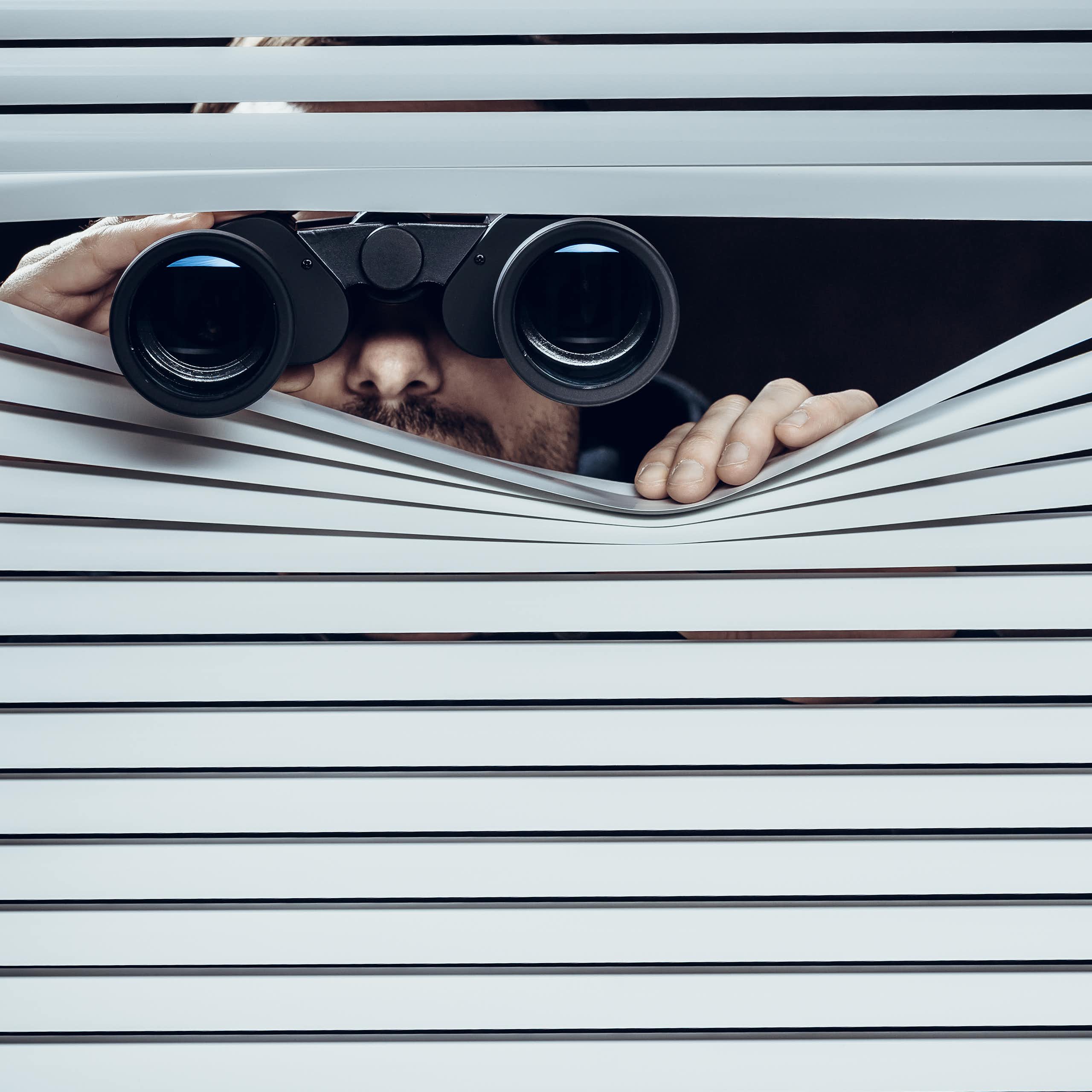 A person using binoculars to secretly look out through a window with blinds in it.