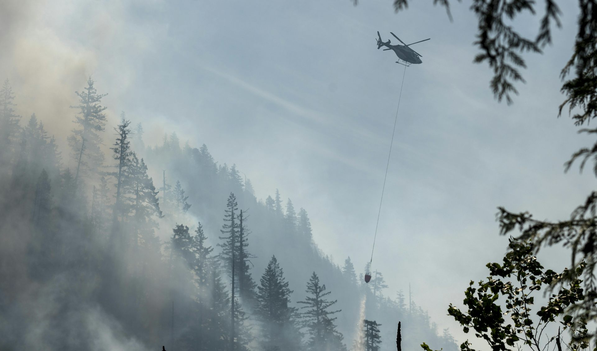 A helicopter drops water on a hilly, smoking forest.
