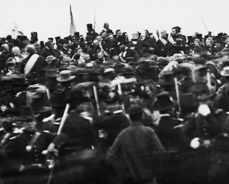An antique photo of a crowd of soldiers and civilians listening to someone talking in the middle.