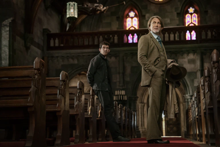 A priest and a well-dressed man in a suit standing amongst the pews of a church.