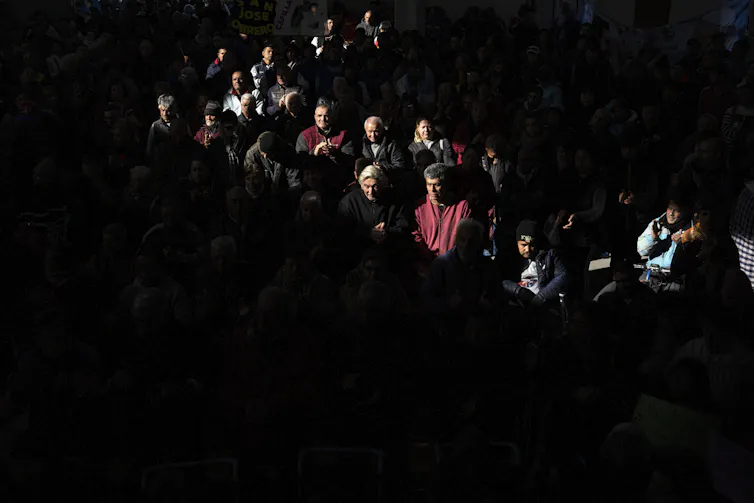 A spotlight shines on a zigzag row of people wearing jackets, with the rest of the crowd hidden in the dark.