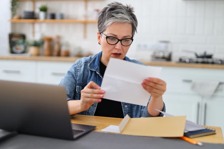 Middle-aged woman sitting at kitchen table, reading letter, with confused expression