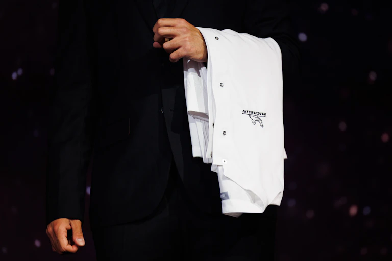 Person wearing black suit holds a white chef's jacket draped over their left arm