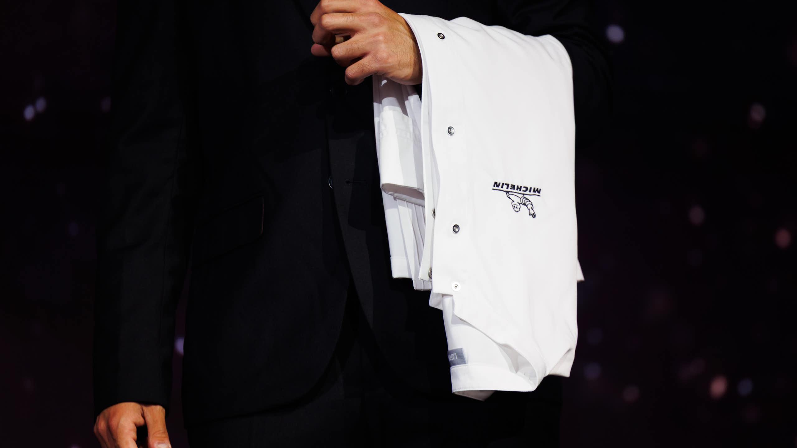 Person wearing black suit holds a white chef's jacket draped over their left arm