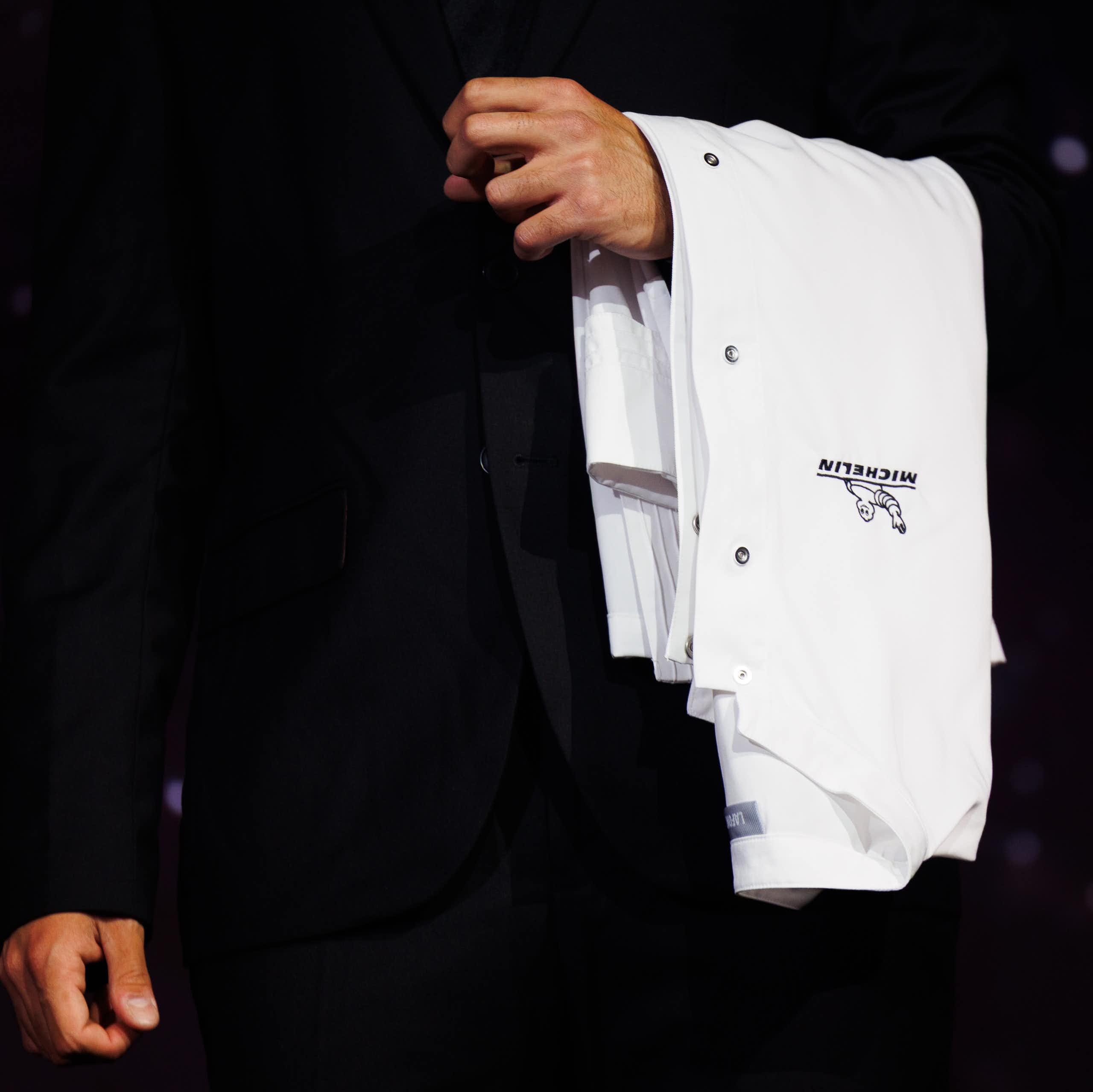 Person wearing black suit holds a white chef's jacket draped over their left arm