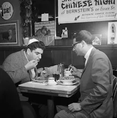 A black and white photograph of two men in suit jackets and Jewish head coverings seated at a table with signs on the wall.