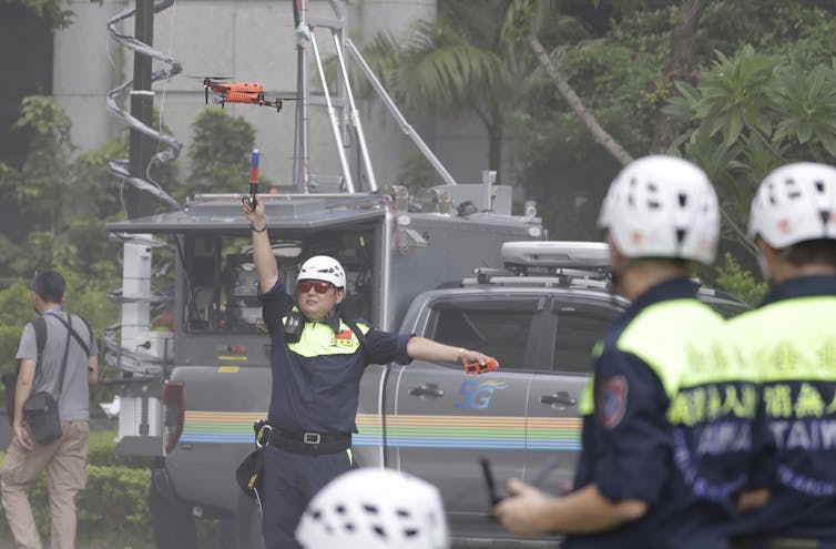 A man in a police uniform and wearing a white helmet operates a drone on a city street.