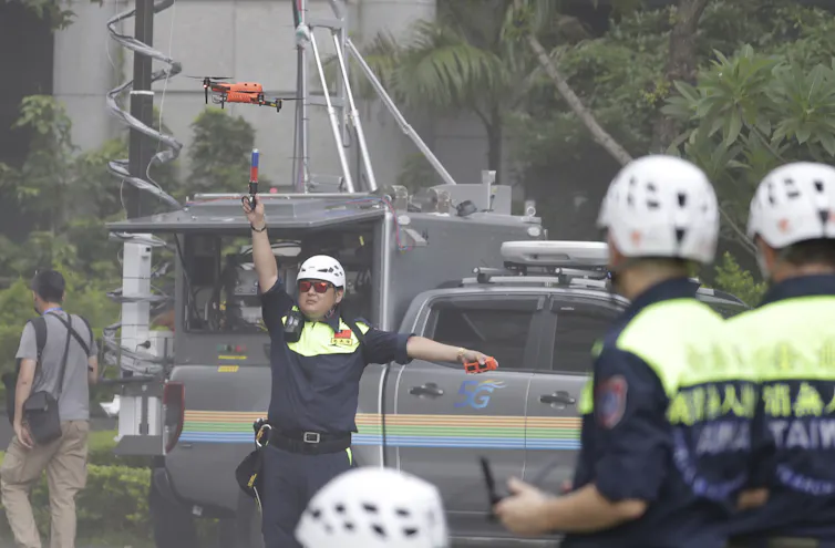 A man in a police uniform and wearing a white helmet operates a drone on a city street.