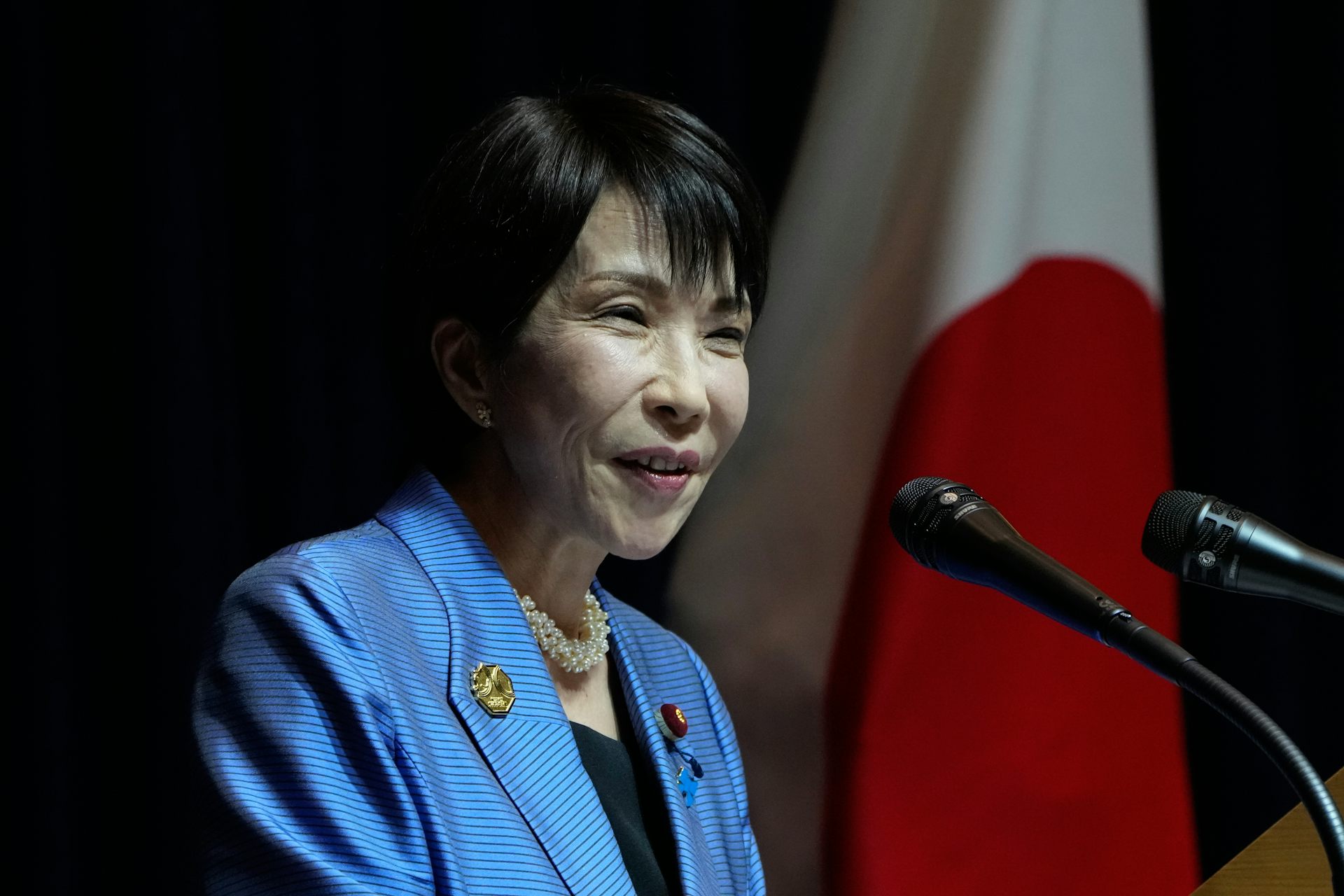 A smiling Asian woman with short dark hair in a periwinkle blue jacket behind two microphones.