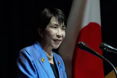 A smiling Asian woman with short dark hair in a periwinkle blue jacket behind two microphones.