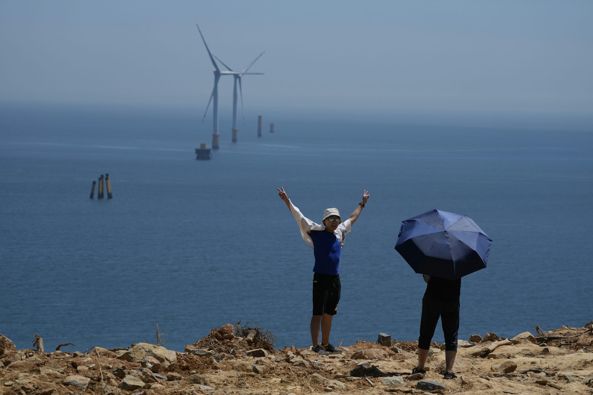 A man poses with his arms over his head on a cliff with the blue sea and wind turbines behind him.