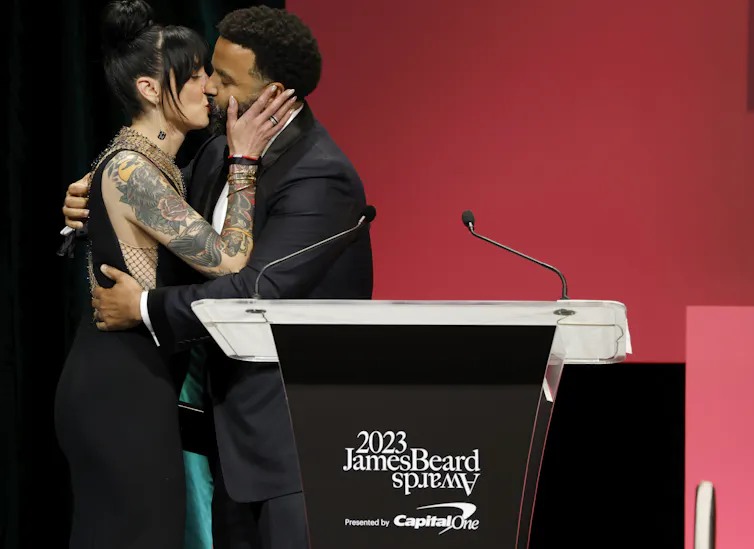Man and women in formalwear kiss on a stage behind a podium that says '2023 James Beard Awards'