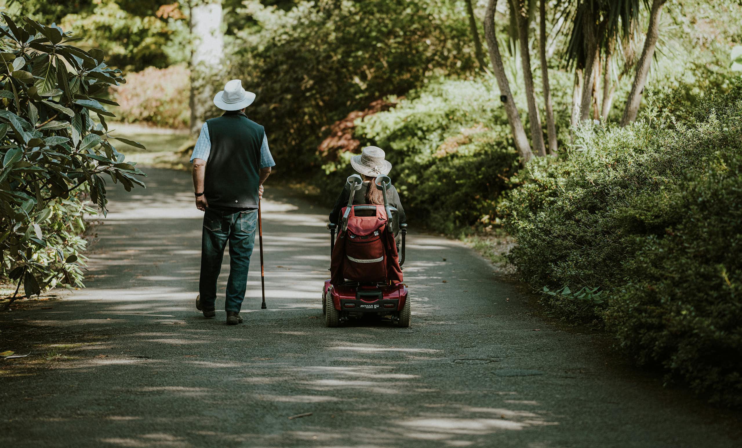 Two people on a path bordered by greenery; one using a cane and the other using a wheelchair