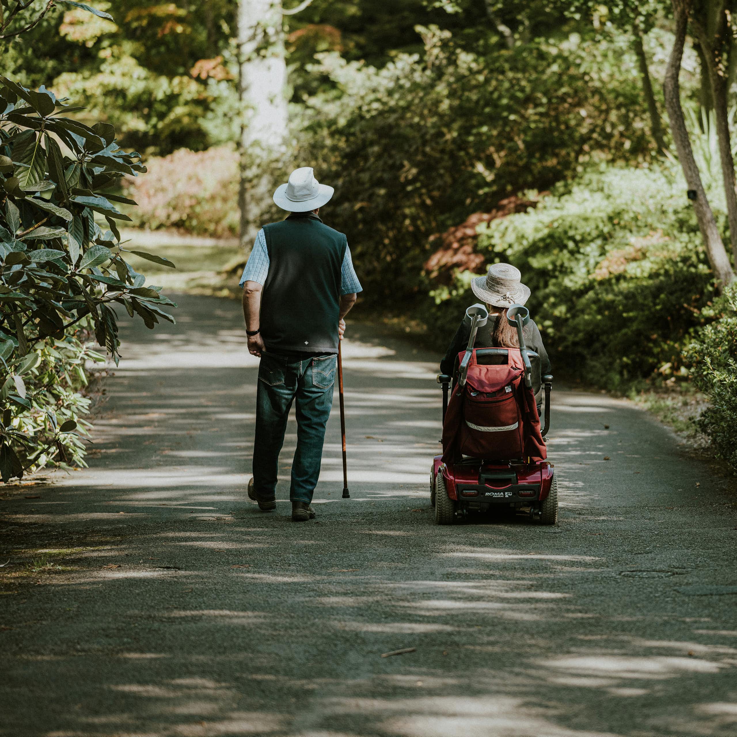 Two people on a path bordered by greenery; one using a cane and the other using a wheelchair