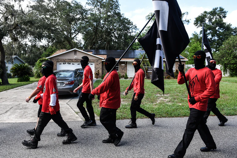 a group of men in red t-shirts and black pants carrying black flags with a white symbols walk down a suburban street