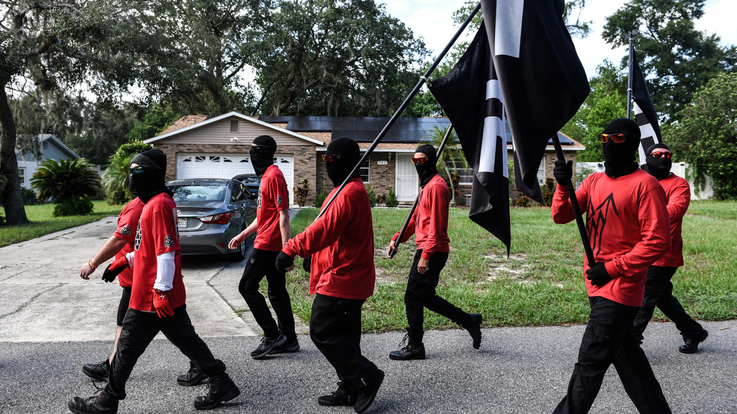 a group of men in red t-shirts and black pants carrying black flags with a white symbols walk down a suburban street
