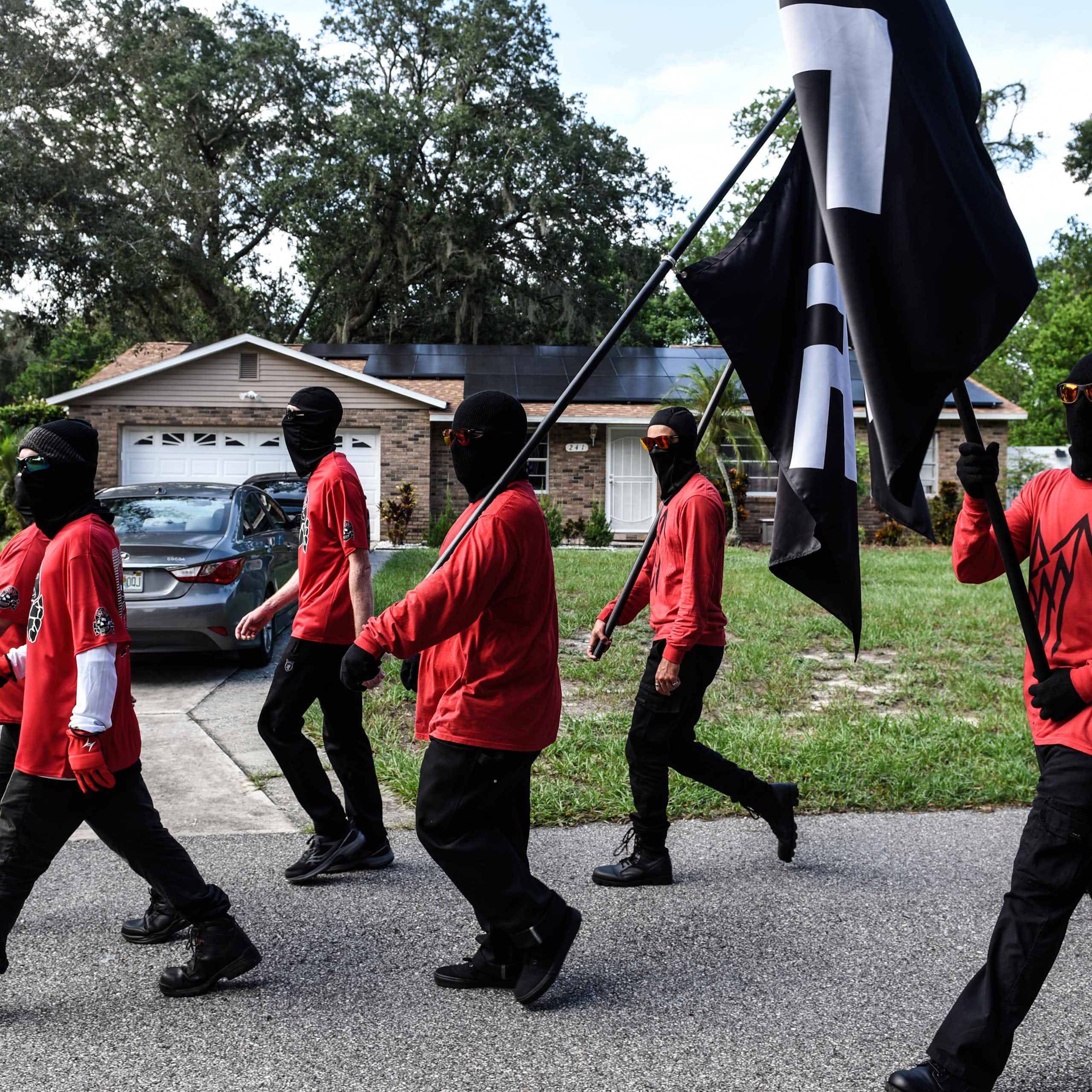 a group of men in red t-shirts and black pants carrying black flags with a white symbols walk down a suburban street