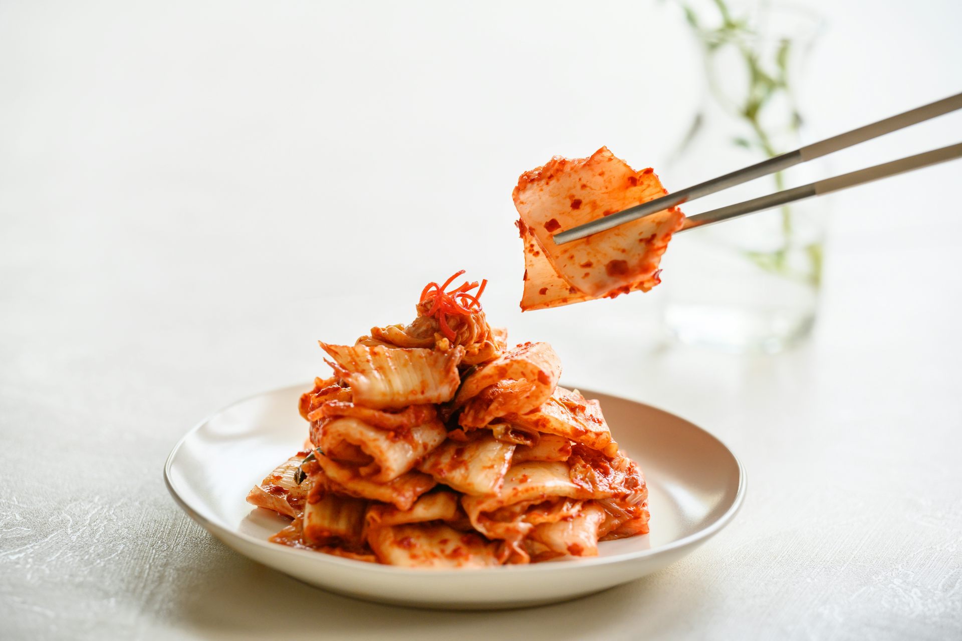 A pair of metal chopsticks picks up kimchi from a white bowl.