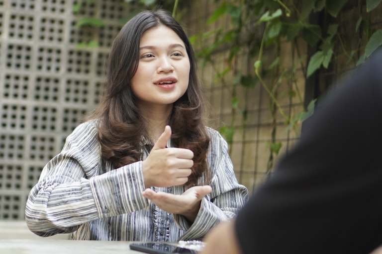 young woman seated talking holds one hand with a thumb up over her other hand