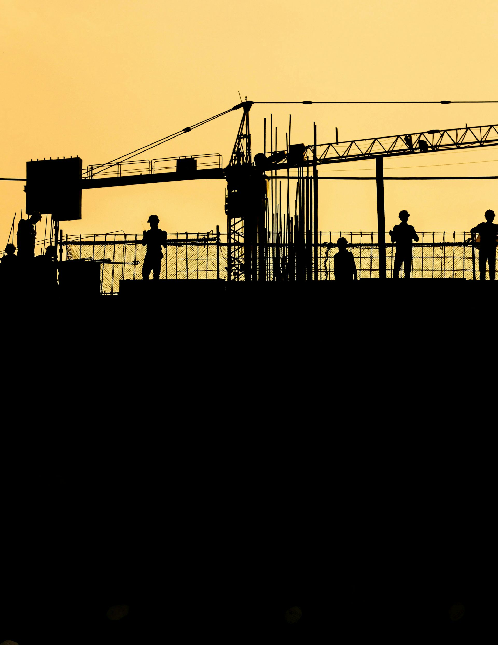 Silhouetted workers in hard hats on a site against an orange sky.