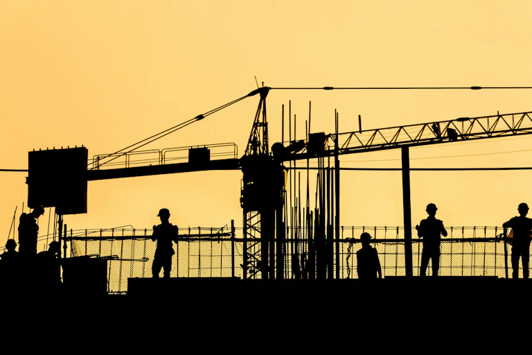 Silhouetted workers in hard hats on a site against an orange sky.