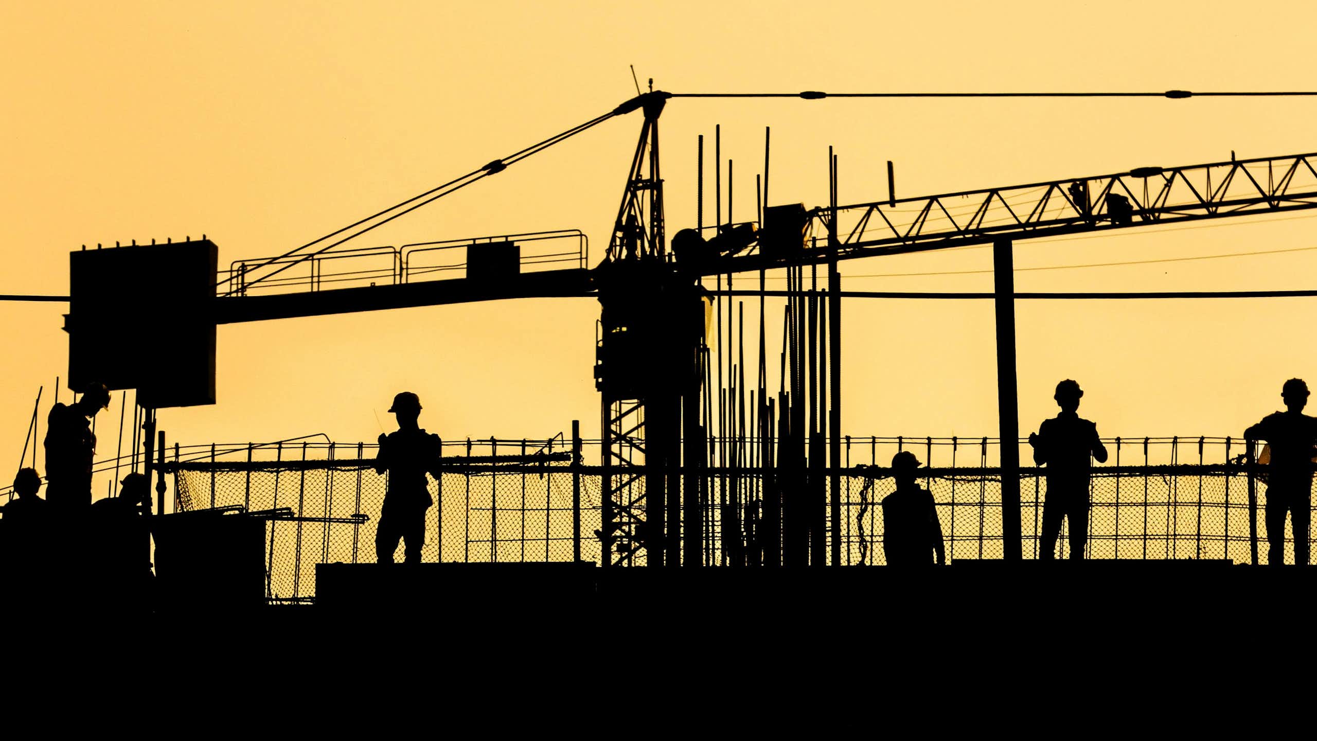 Silhouetted workers in hard hats on a site against an orange sky.