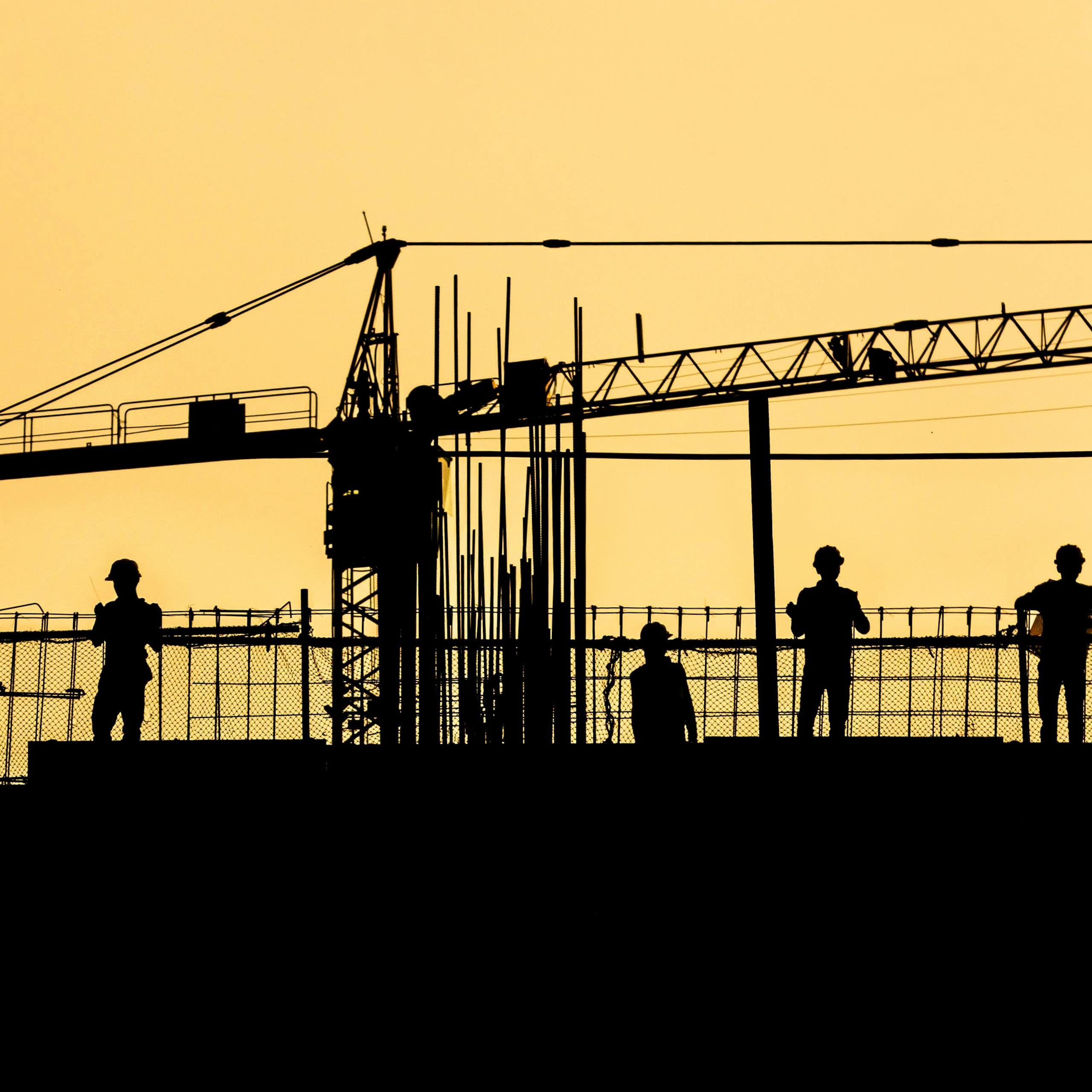Silhouetted workers in hard hats on a site against an orange sky.