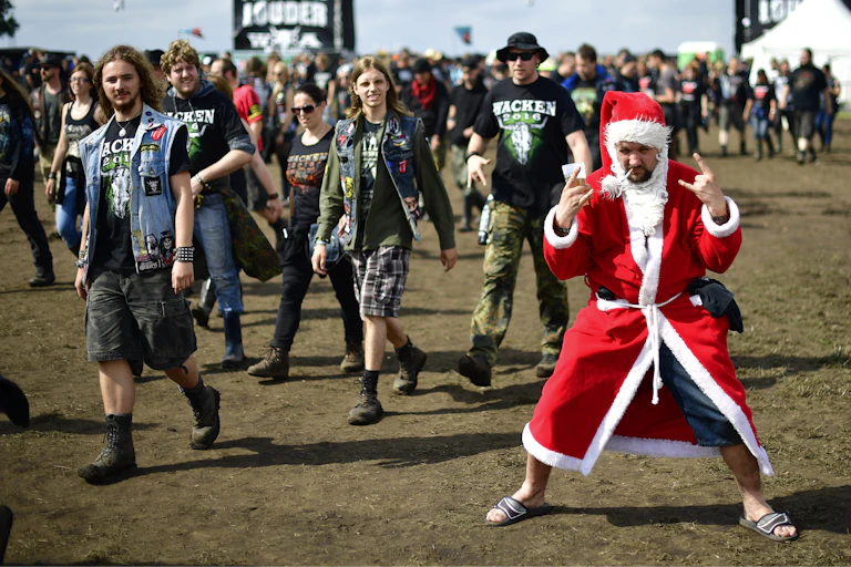 A young man dressed in a Santa suit smokes a cigarette and makes devil horn signs with his hands at a music festival.