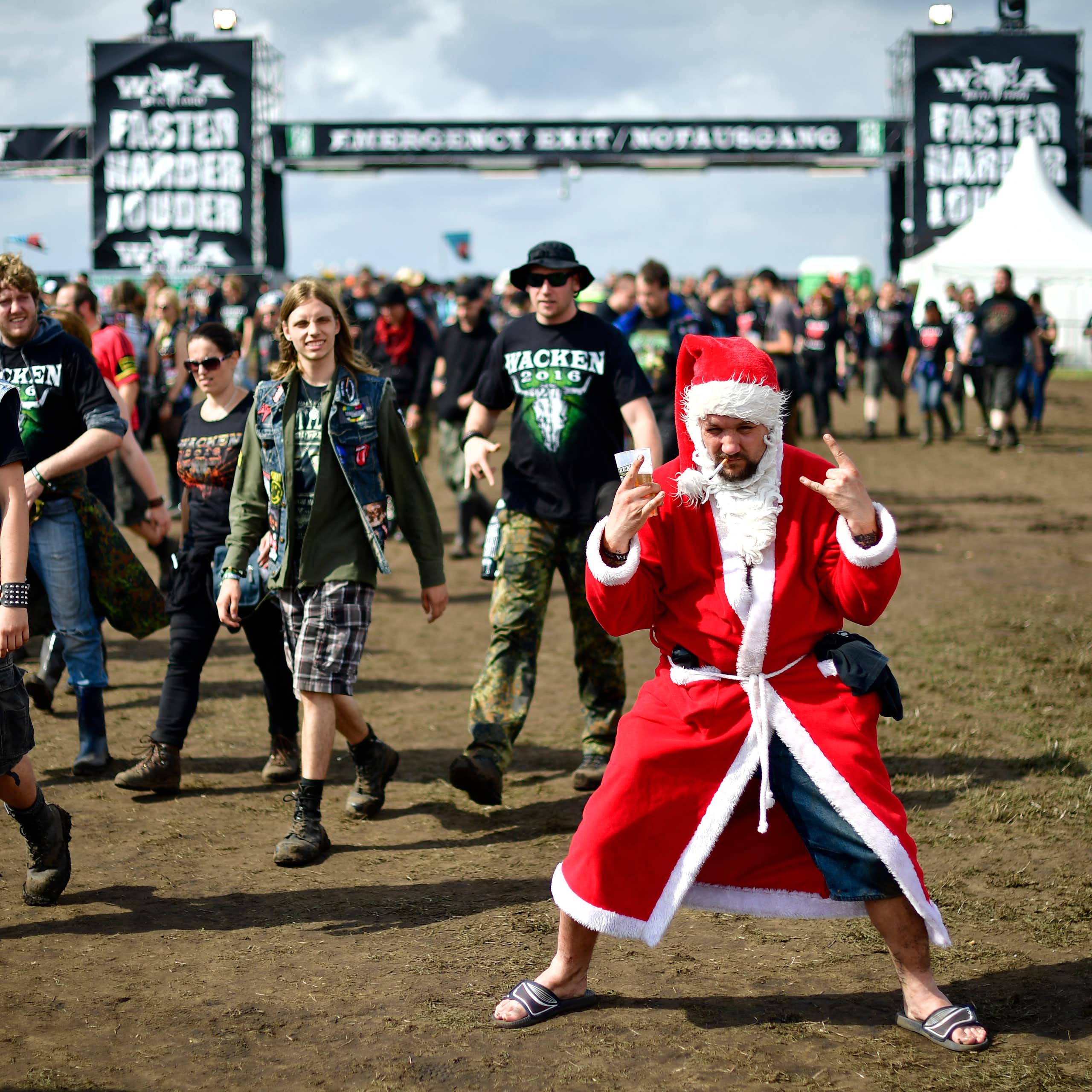 A young man dressed in a Santa suit smokes a cigarette and makes devil horn signs with his hands at a music festival.