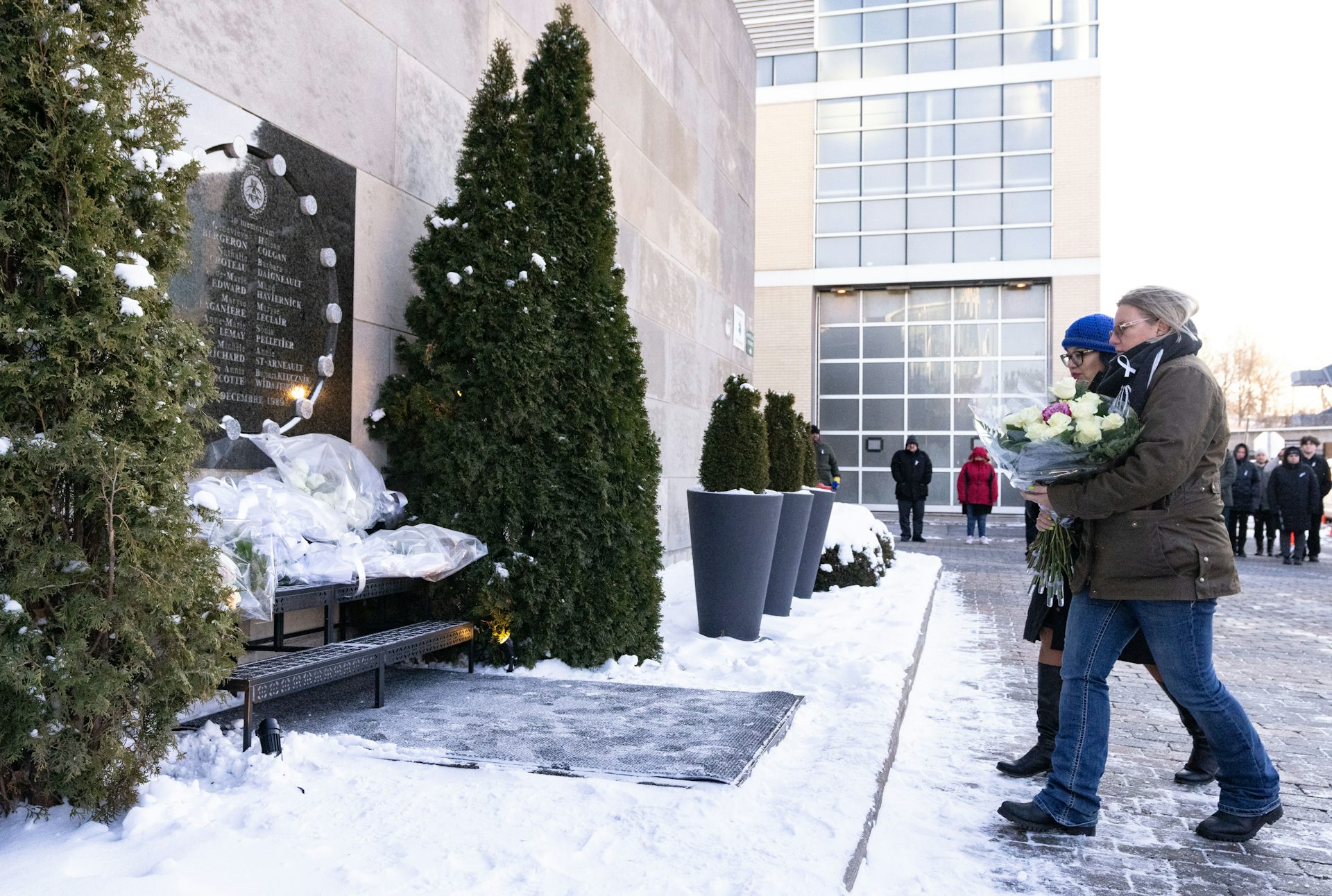 People approach a snowy outdoor memorial, coniferous trees on either side, carrying bouquets of white flowers.