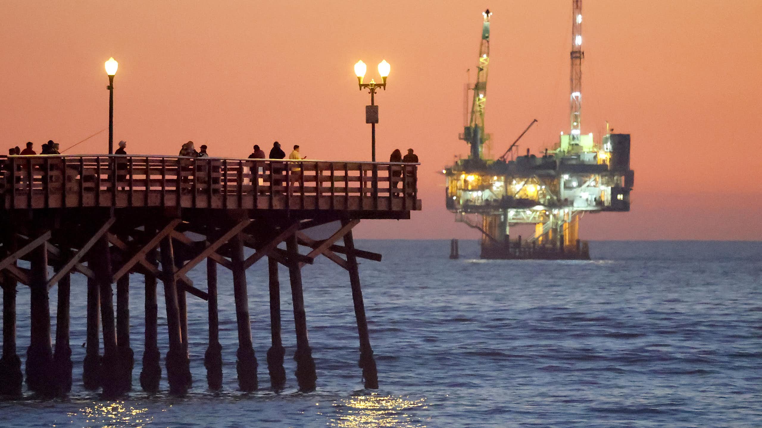 A long wooden pier stretches into the ocean, where a brightly lit industrial platform rises from the surface of the sea.