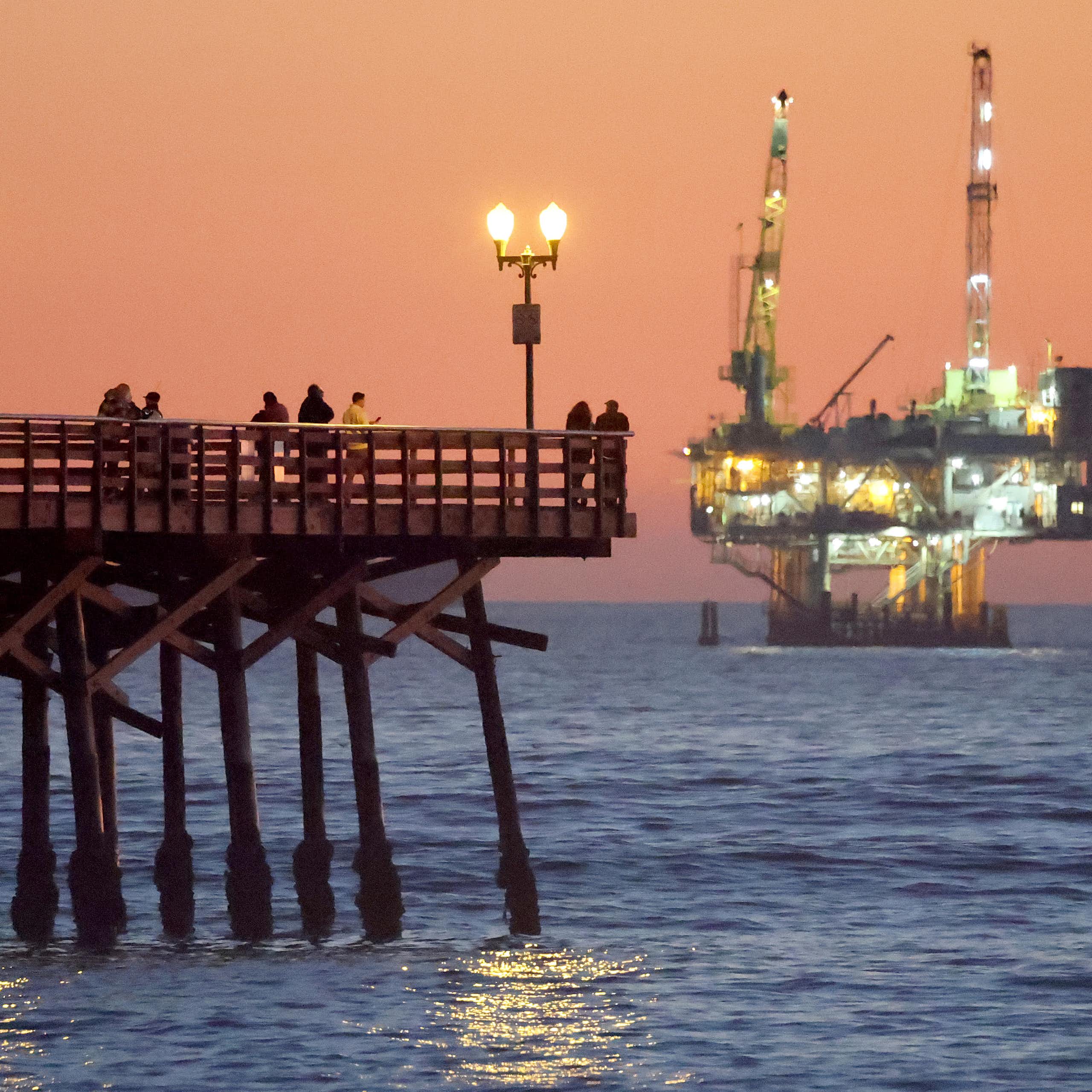 A long wooden pier stretches into the ocean, where a brightly lit industrial platform rises from the surface of the sea.