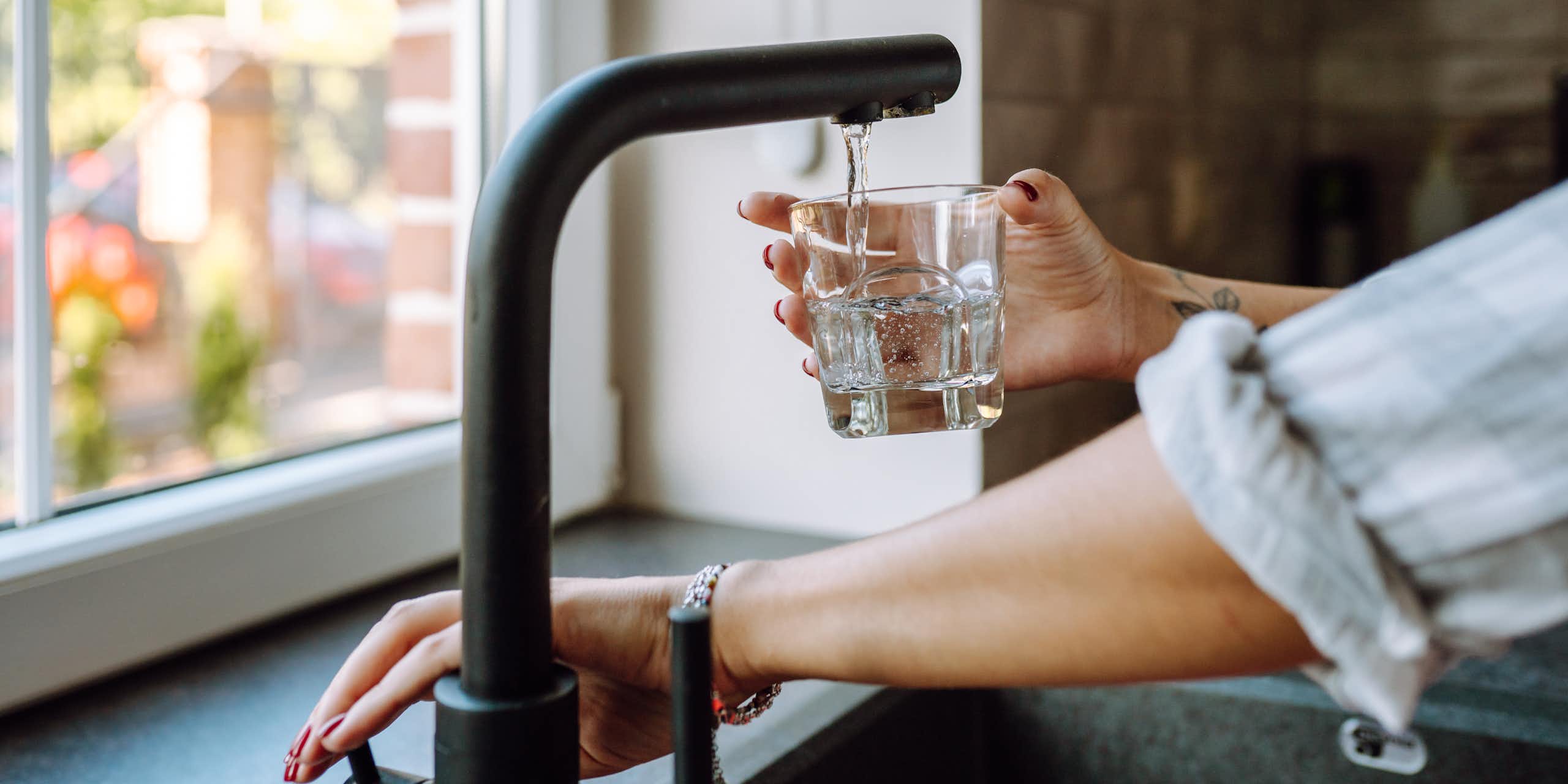 A woman's hands fill a glass with water. She has a tattoo on her wrist and rolled up sleeves.