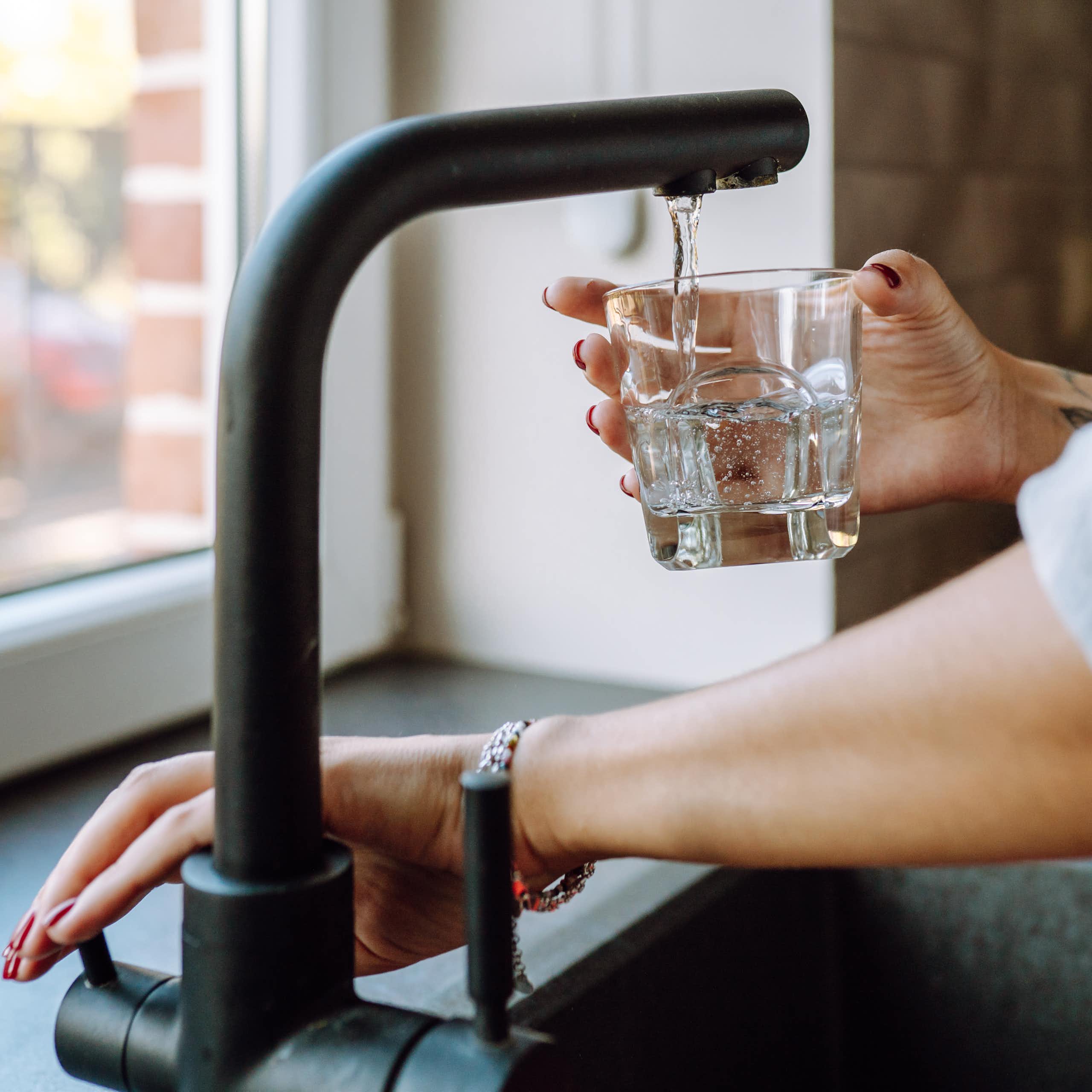 A woman's hands fill a glass with water. She has a tattoo on her wrist and rolled up sleeves.