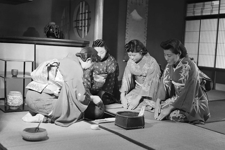 Women seated on a tatami mat wearing kimonos; one serves Japanese tea while the others wait.
