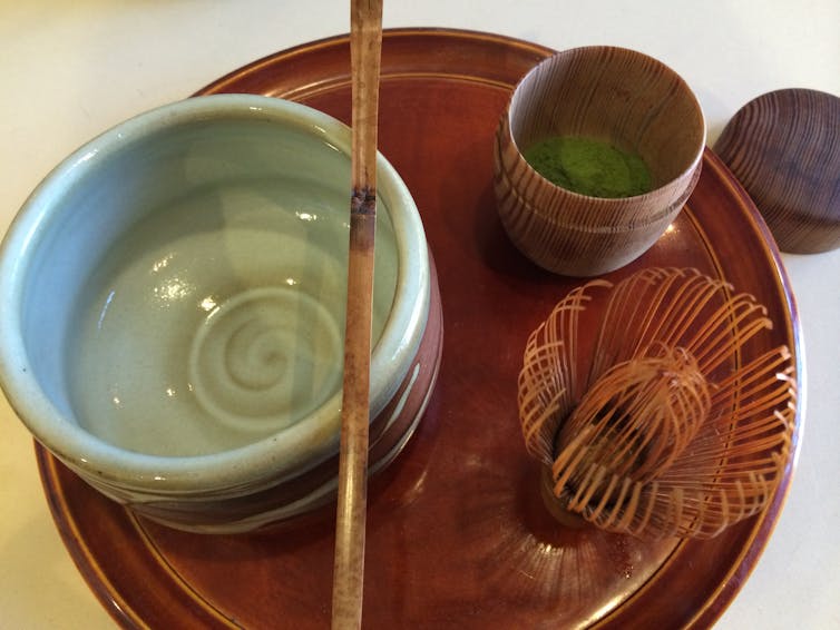 A wooden plate holding a blue ceramic bowl, a whisk, a cup and a spoon.