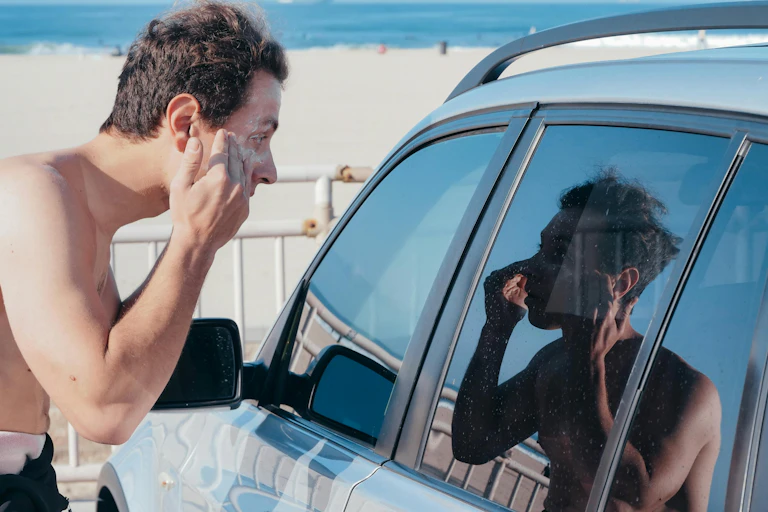 Man applies sunscreen at beach while looking into the car window reflection.