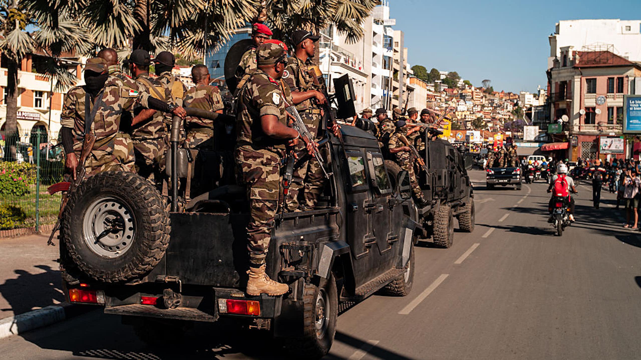 Armed men standing on the back of a truck that is part of a convoy driving through a street
