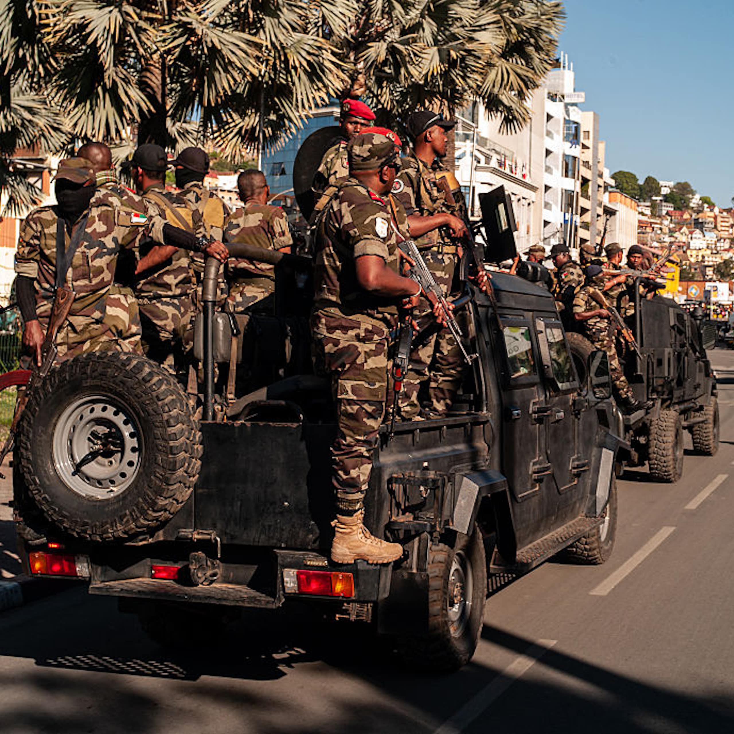 Armed men standing on the back of a truck that is part of a convoy driving through a street