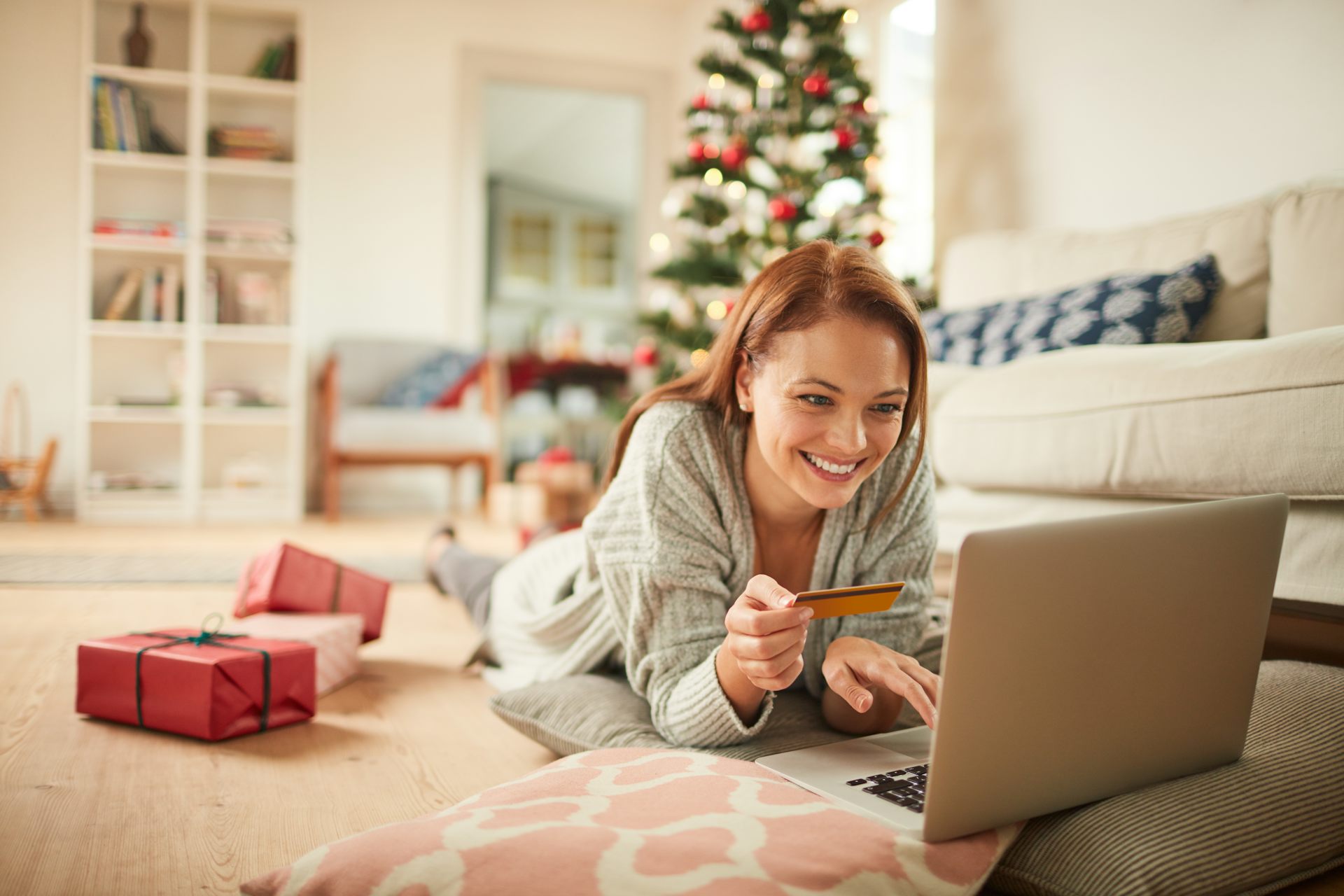 Woman lying on living room floor with laptop holding a bank card, Christmas tree in background