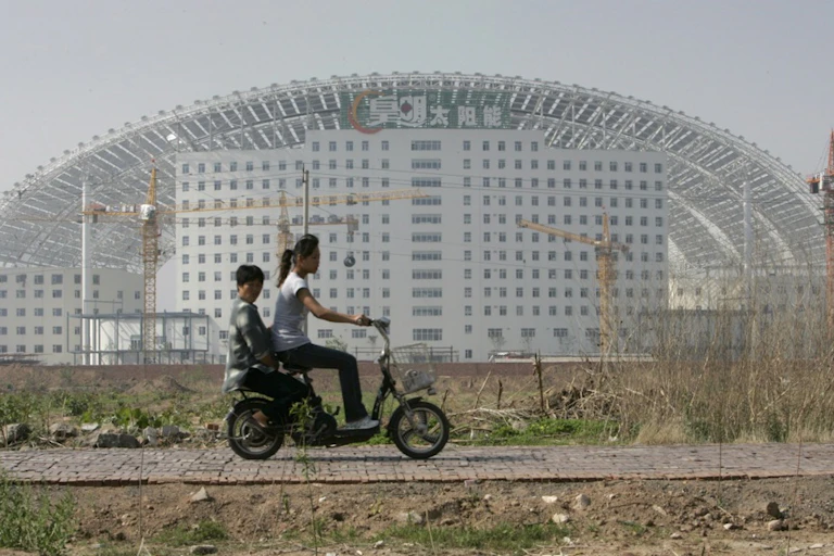 Two people riding an electric scooter past a large building.