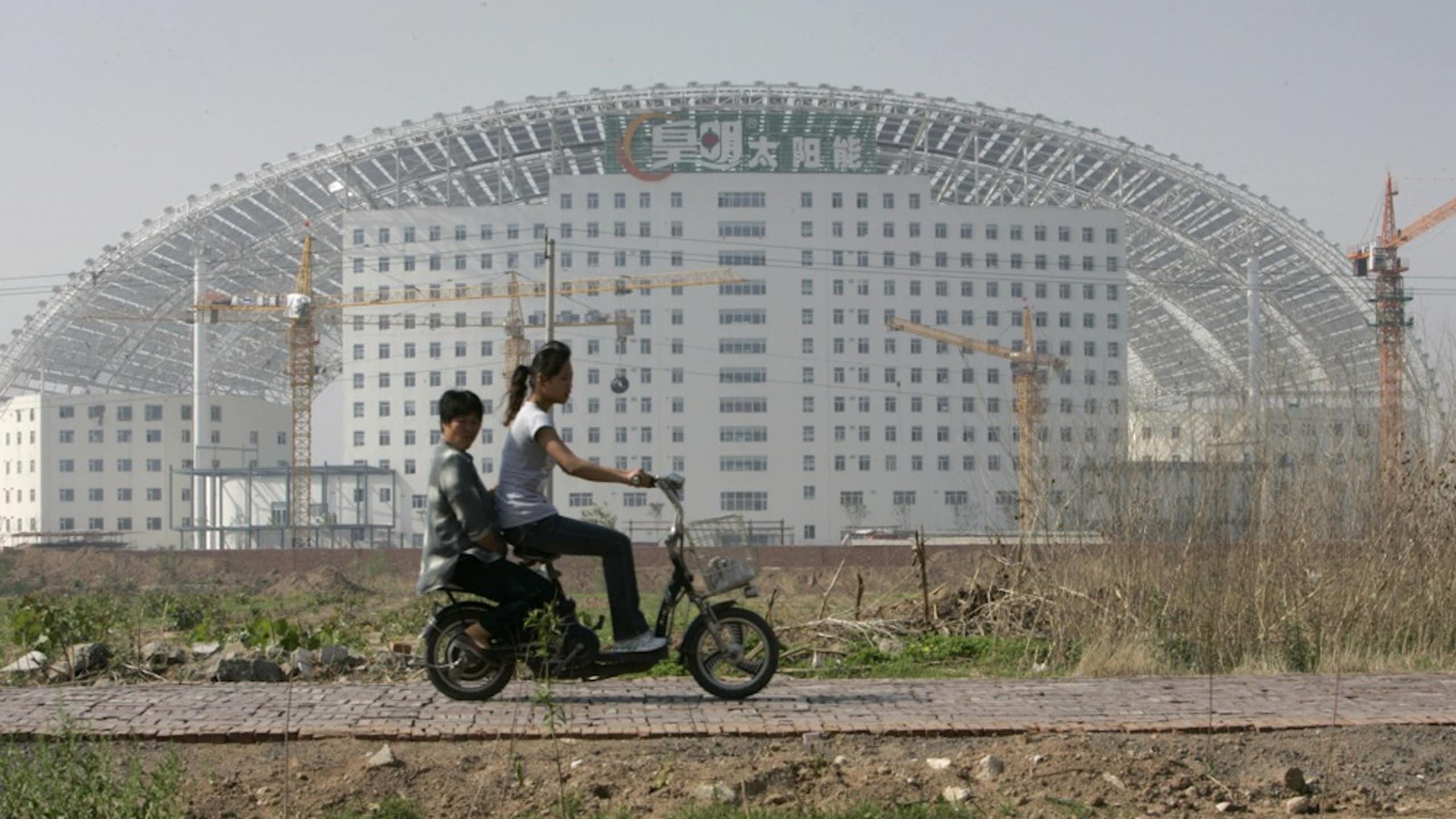 Two people riding an electric scooter past a large building.