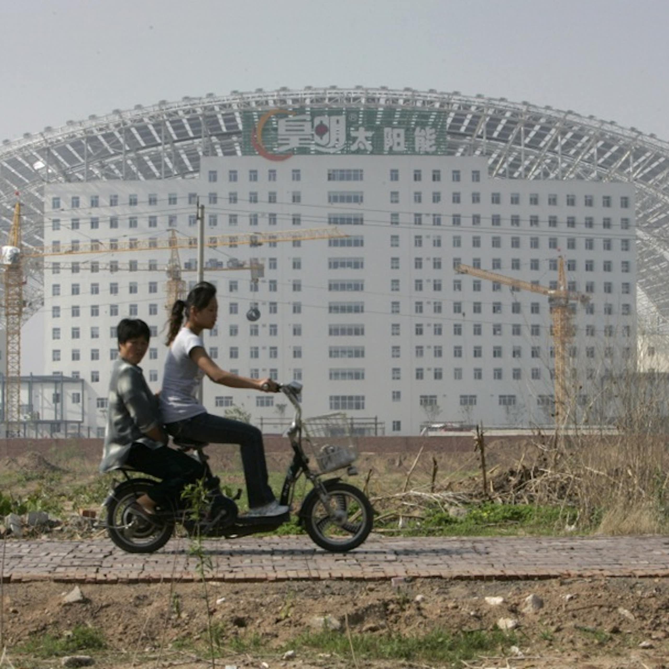 Two people riding an electric scooter past a large building.