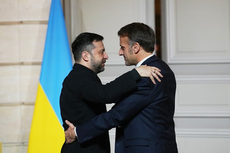 Two dark-haired men hug in front of a blue and yellow flag.