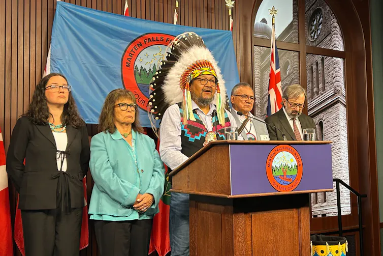 Un hombre con un tocado de plumas indĆgenas habla en un podio, detrĆ”s de Ć©l hay otras personas.