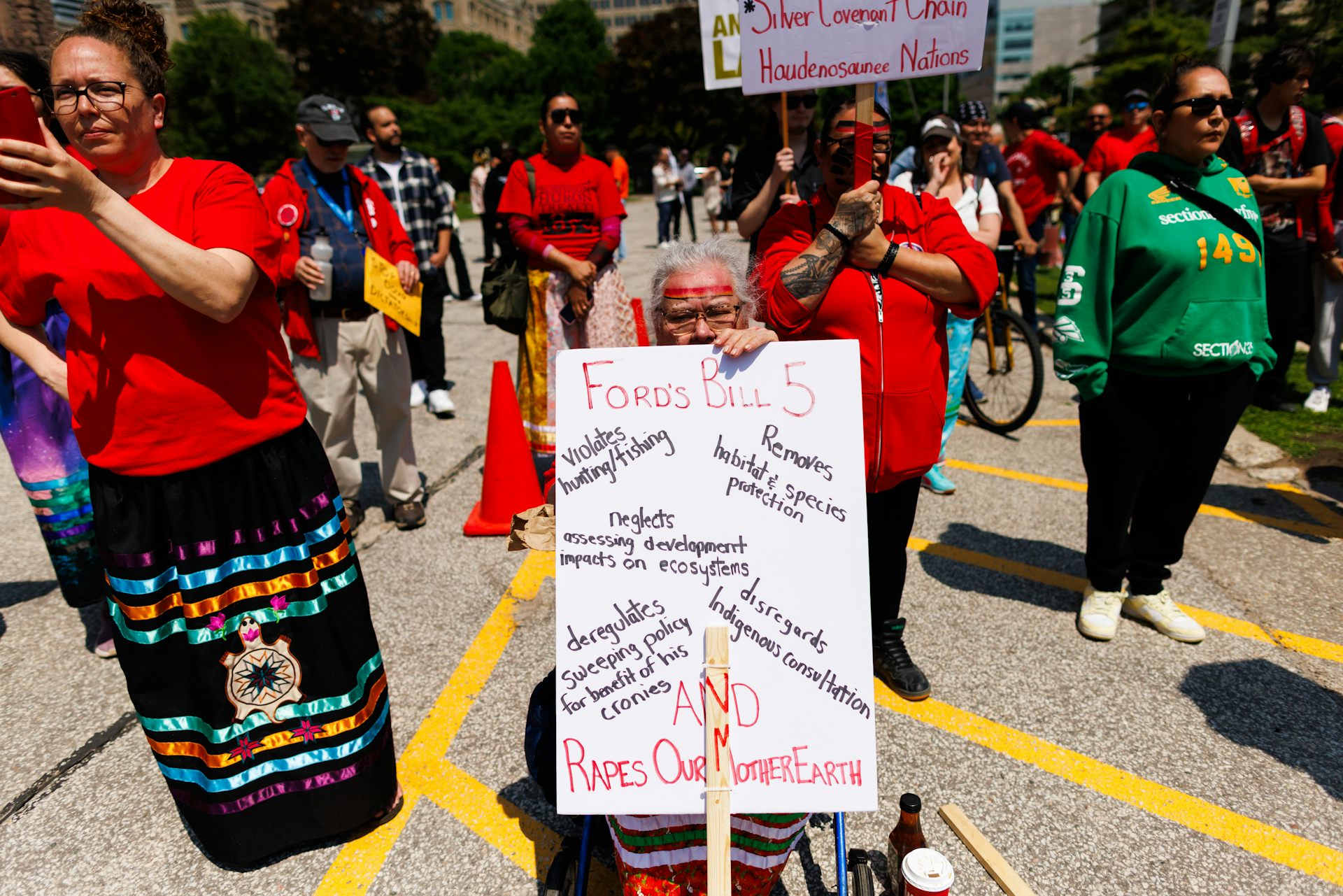 Una anciana en la protesta lleva un cartel criticando la Ley 5. Otras personas con camisetas rojas están paradas a su alrededor.