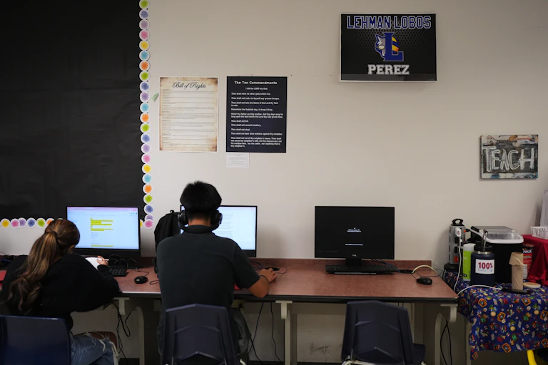 Two teenagers with their backs to the camera work at computers on a long table, pushed up against a white wall.