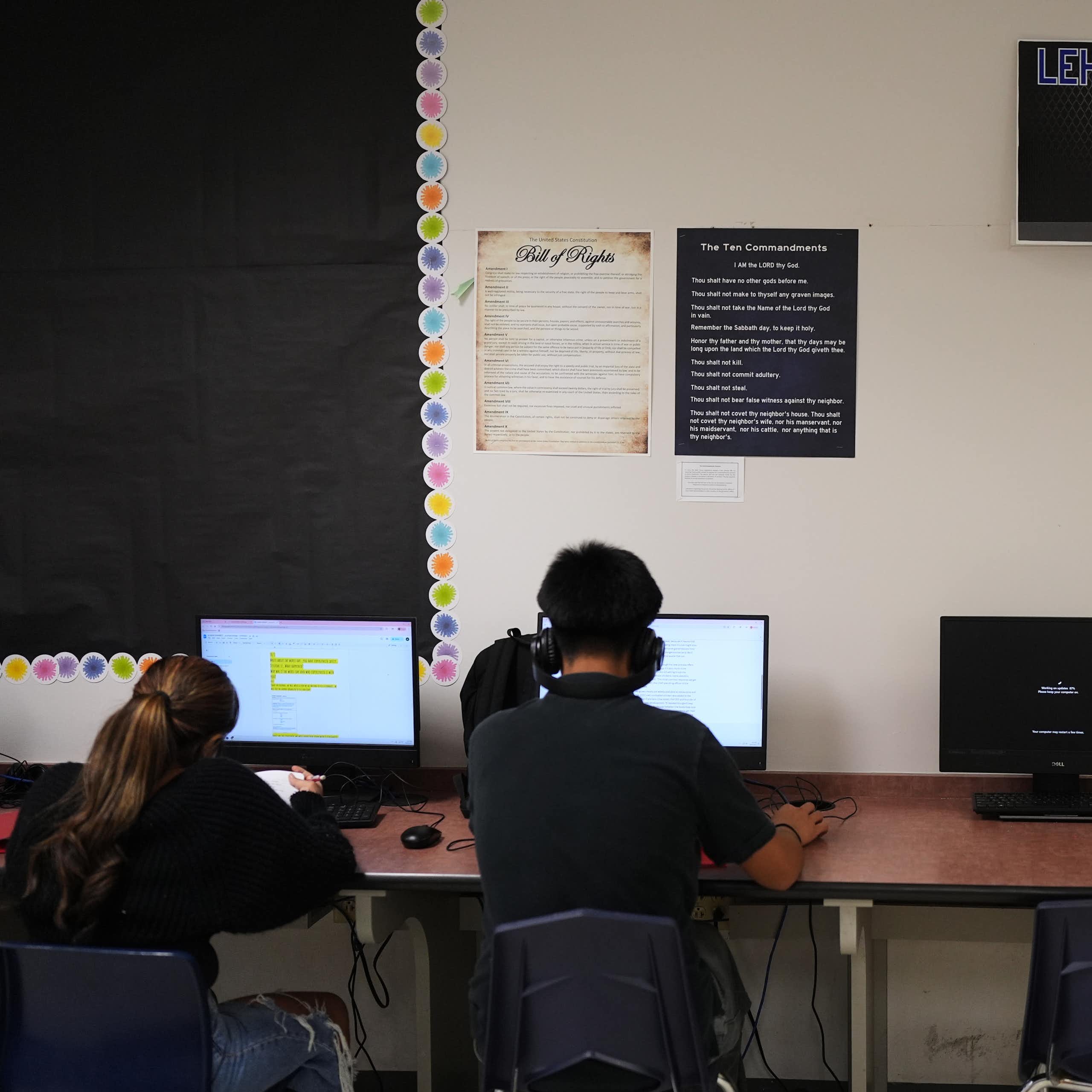 Two teenagers with their backs to the camera work at computers on a long table, pushed up against a white wall.