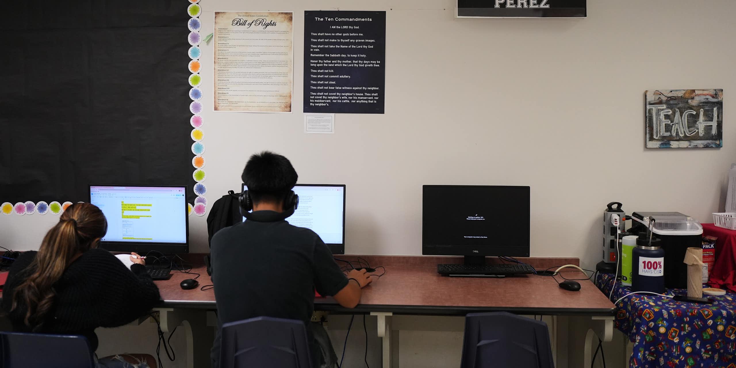 Two teenagers with their backs to the camera work at computers on a long table, pushed up against a white wall.