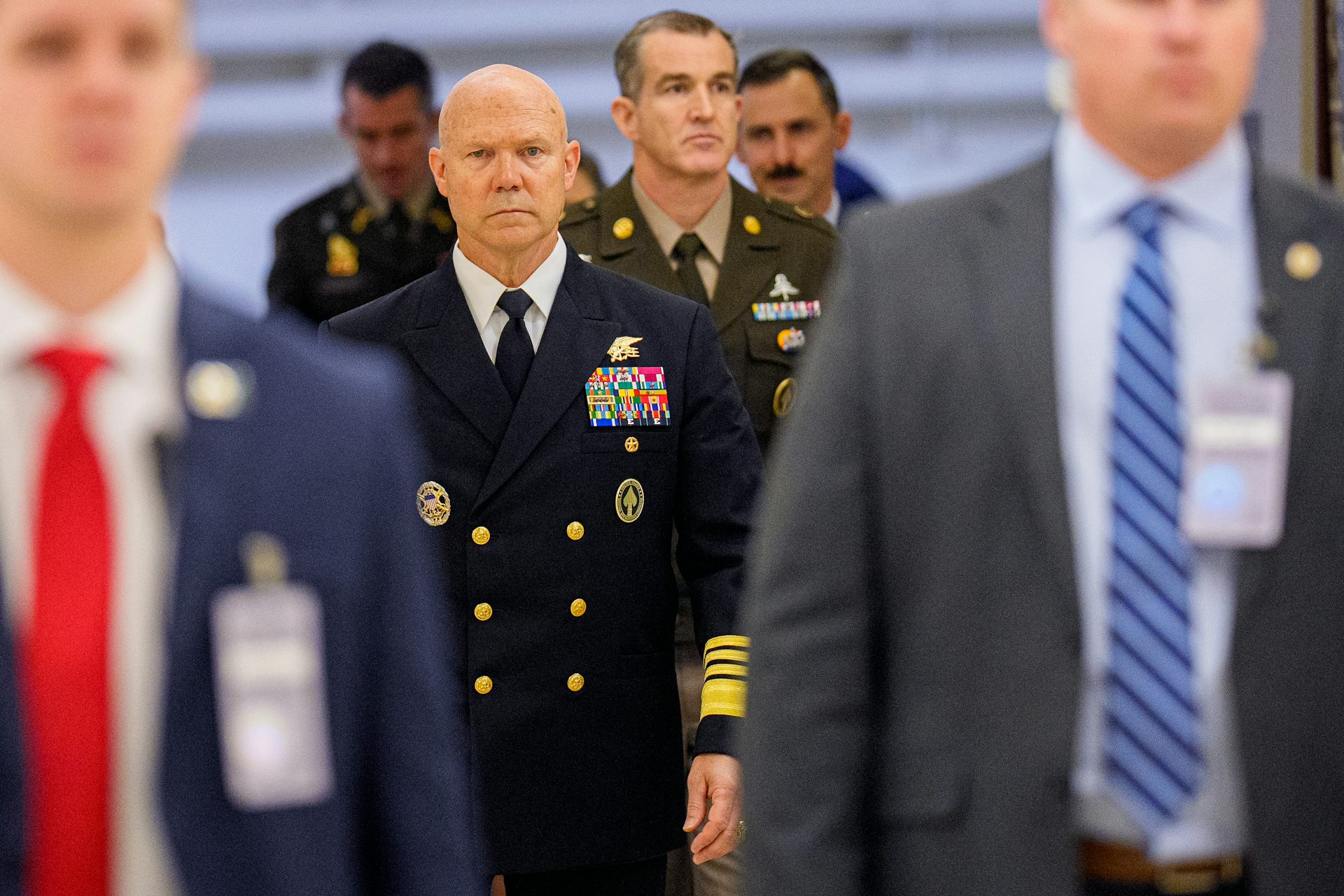 A man in a dark blue military uniform walks among a number of men in suits.